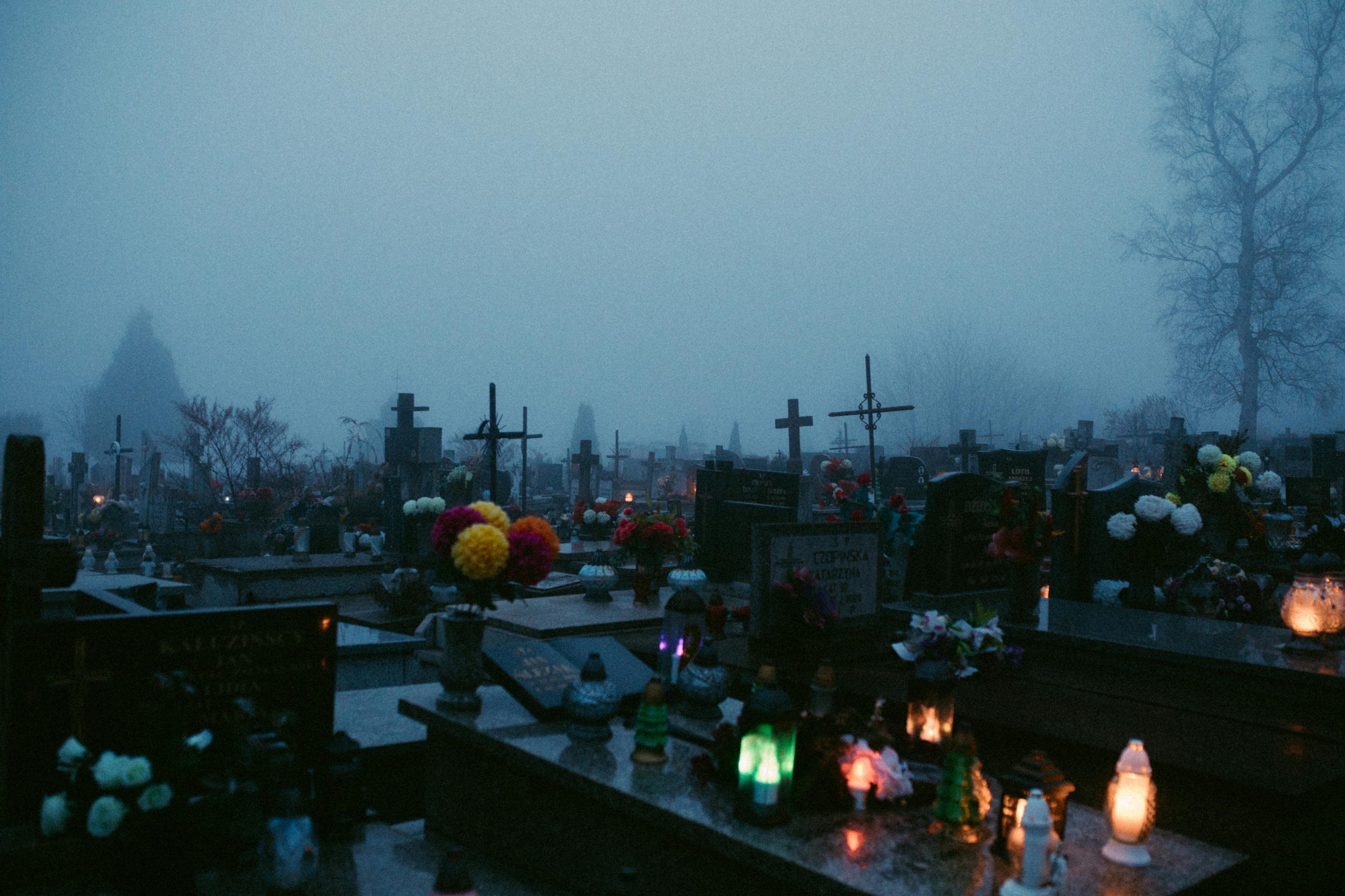 A foggy cemetery at night illuminated by candlelights on the graves, evoking a somber ambiance.